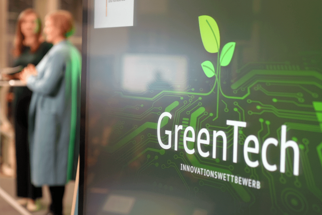 Im Vordergrund das GreenTech Logo aus einer grünen Wand, dahinter zwei Personen, die sich unterhalten