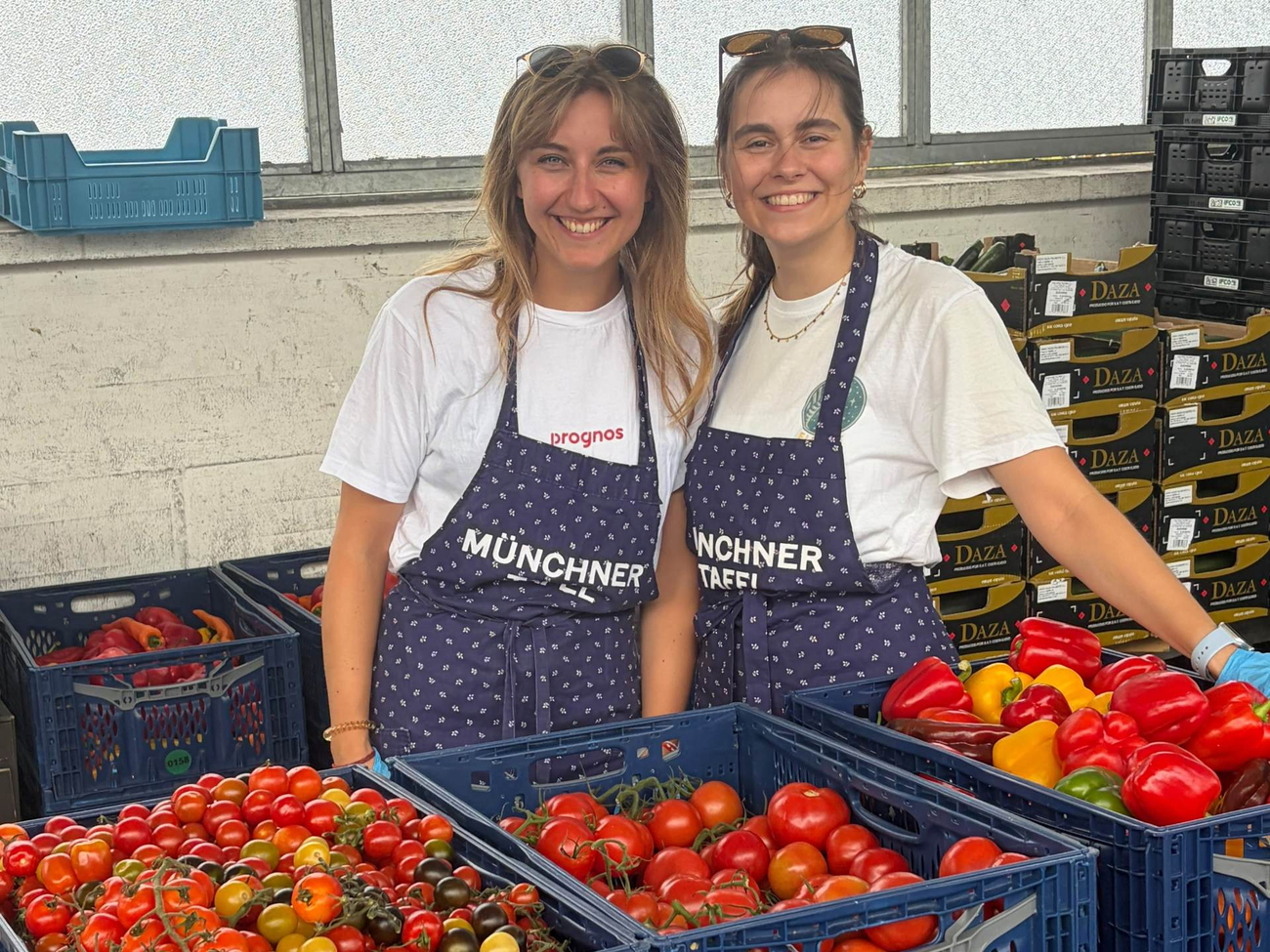Zwei lächelnde Frauen in Schürzen stehen hinter Kisten mit Tomaten und Paprika.