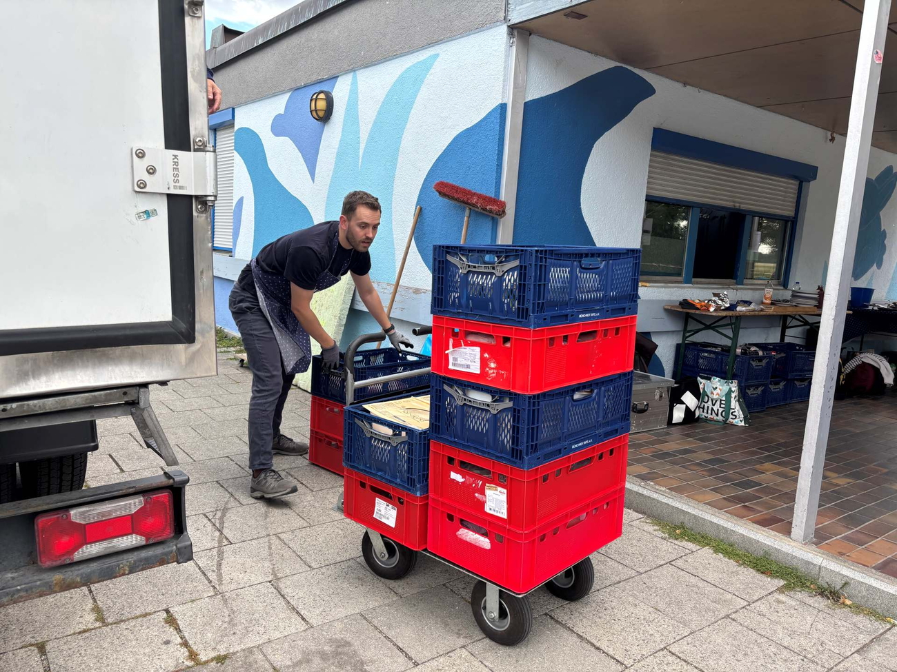 Mann schiebt roten Stapel Kisten auf Rollwagen vor Werkstatt, Transporter daneben
