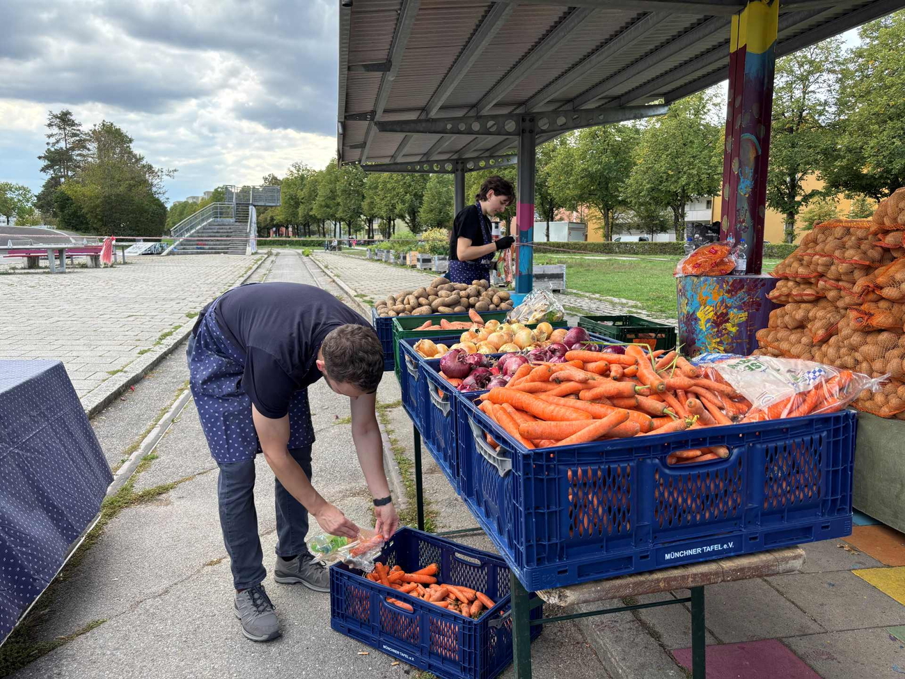 Mann beugt sich über Kisten mit Karotten am Marktstand, im Hintergrund weitere Ware.
