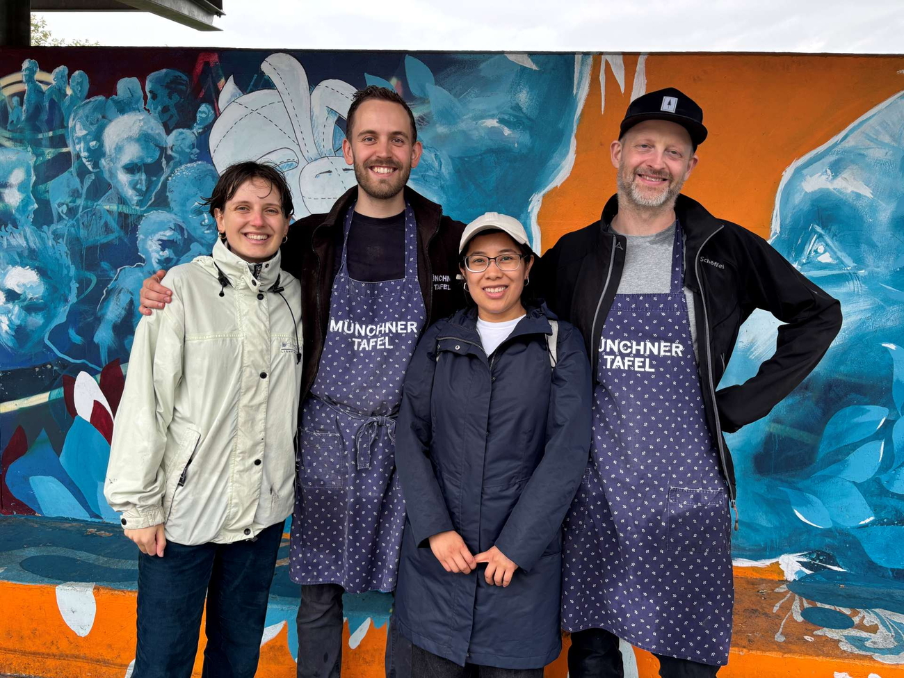 Vier Personen in Schürzen vor blauem Kunstwerk, lächelnd vor orangefarbener Wand.