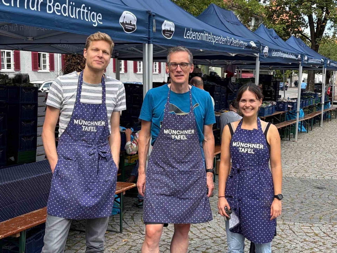 Drei lächelnde Personen in blauen Schürzen auf Marktstand mit Bierbänken.