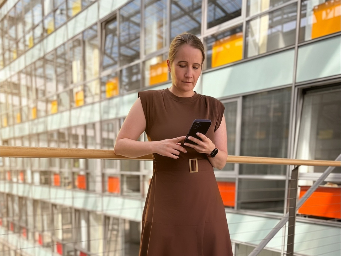 Frau in braunem Kleid liest auf Smartphone in modernem Gebäude mit Glasfassade