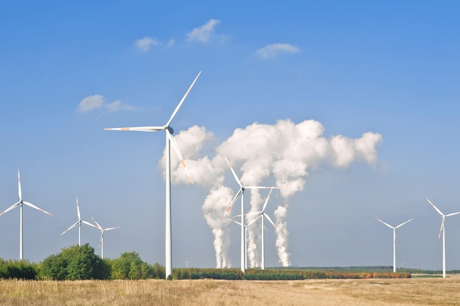 Blick übers Feld. Im Hintergrund zu sehen Windräder und eine Fabrik mit Abgasen