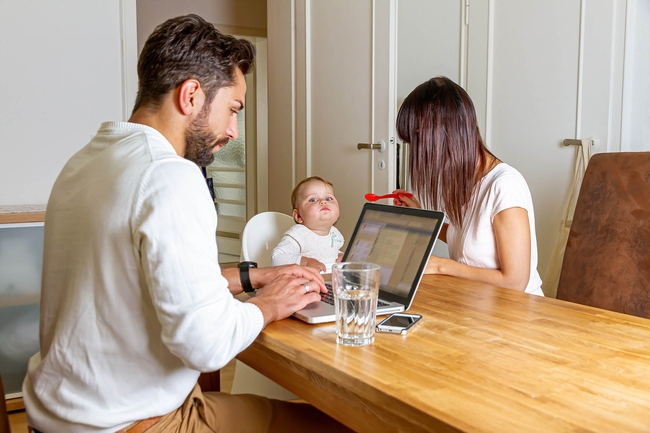 Vater Mutter und Knd sitzen am Küchentisch. Vater arbeitet, Mitter füttert das Kind 