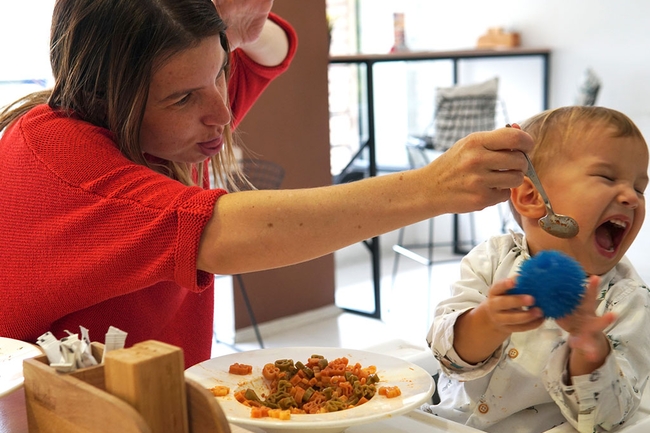 Weinendes Kind weigert sich, zu essen, während Mutter versucht, das Kleinkind durch Tricks zu täuschen