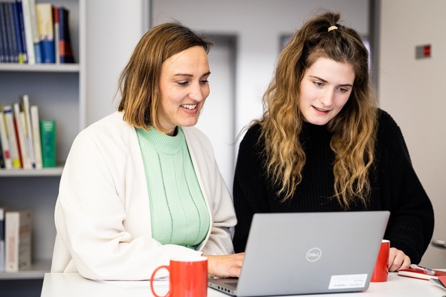 Zwei Frauen arbeiten zusammen am Stehtisch, im Hintergrund Bücher