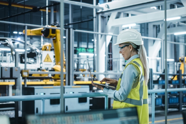 Frau mit Wanrweste und Helm steht mit Laptop in einer Produktionshalle, in der Autos von Roboterarmen gefertigt werden