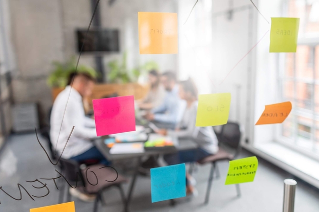 eine Gruppe von Menschen sitzt zusammen in einem Büroraum. An der Glastür kleben Post-ITs