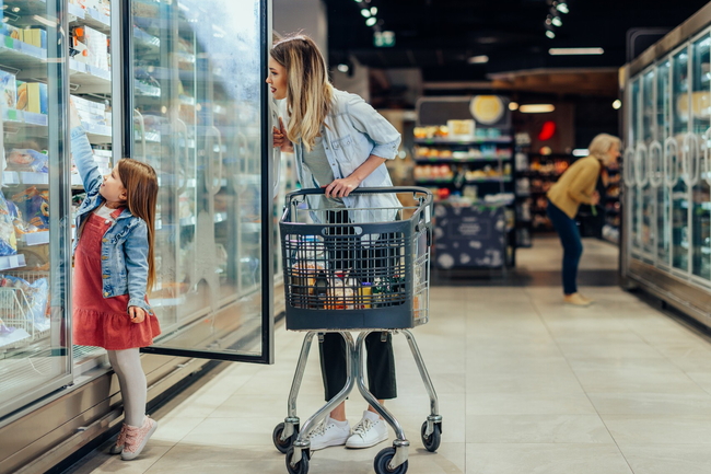 Mädchen blickt im Supermarkt in das offene Tiefkühlregal, Mutter mit Einkaufswagen im Gang.