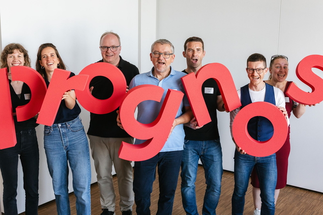Prognos Mitarbeitende halten rote Buchstaben mit dem neuen Prognos Logo in der Hand