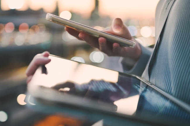 Nahaufnahme von einem Tablet und Handy in der Hand einer Person. Im Hintergrund sieht man eine Stadt.