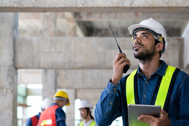 Eine Person auf einer Baustelle mit Warnweste und Helm spricht in ein Walkie-Talkie