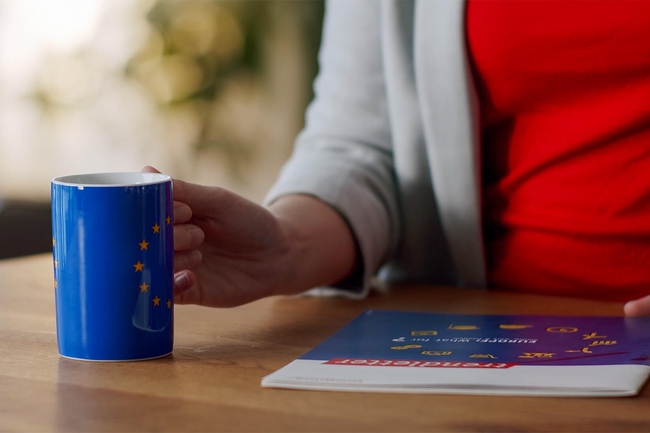 Eine Frau umfasst eine Tasse, auf der die Europaflagge zu sehen ist und schlägt ein Magazin auf