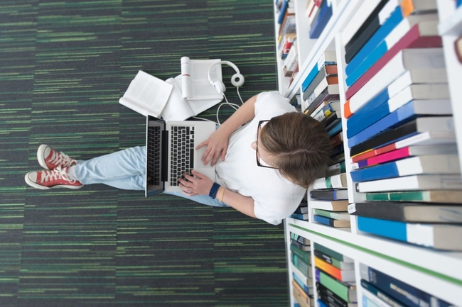 Eine Person sitzt auf dem Boden einer Bibliothek und arbeitet am Laptop