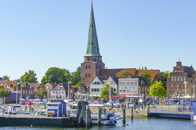 Hafen von Travemünde mit Booten, bunten Häusern und hoher Kirchturmspitze bei Sonnenschein.