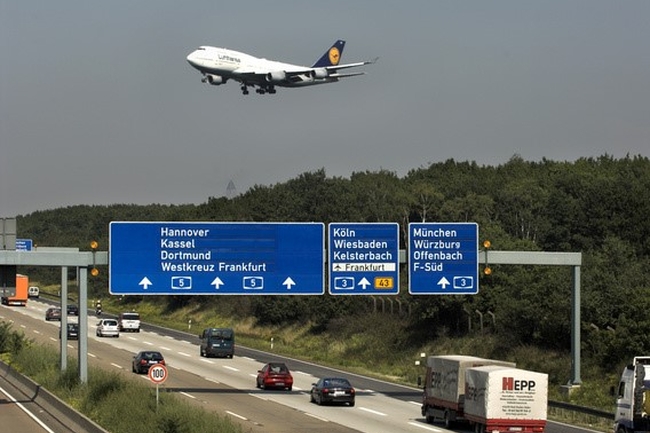 Eine befahrene Autobahn, darüber fliegt ein Flugzeug der Lufthansa