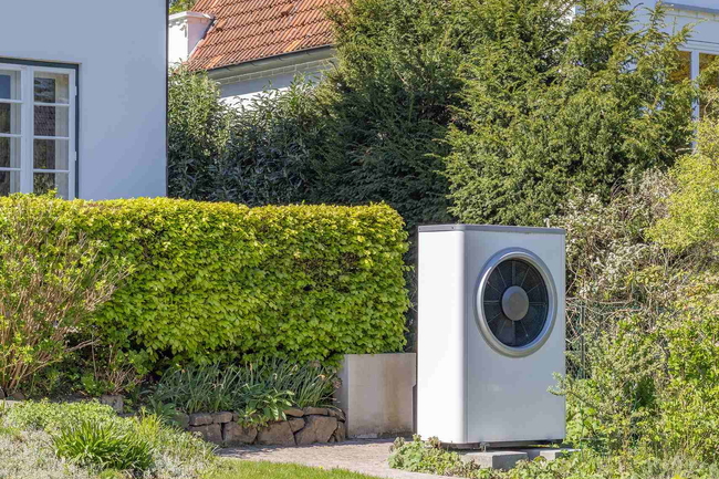 Außeneinheit einer Wärmepumpe im Garten neben Hecke und Hauswand