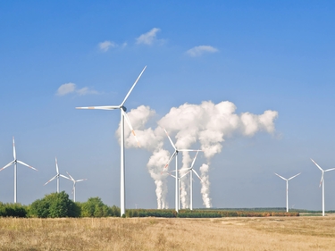 Blick übers Feld. Im Hintergrund zu sehen Windräder und eine Fabrik mit Abgasen