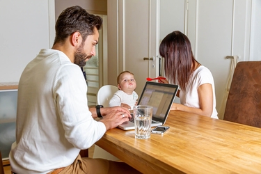 Vater Mutter und Knd sitzen am Küchentisch. Vater arbeitet, Mitter füttert das Kind 