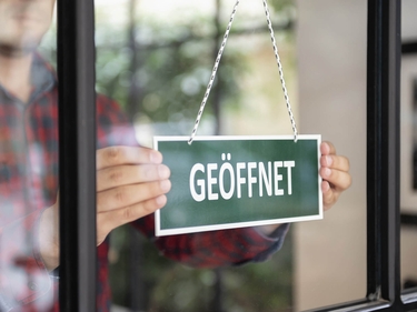 Ein Mann öffnet sein Geschäft. Auf einem grünen Schild steht "Geöffnet"