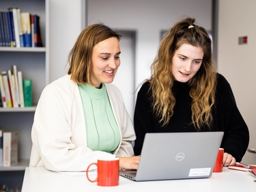 Zwei Frauen arbeiten zusammen am Stehtisch, im Hintergrund Bücher