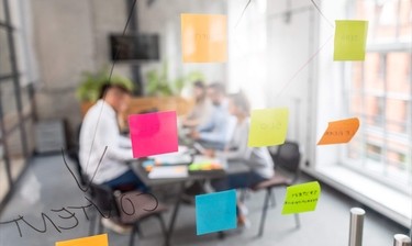 eine Gruppe von Menschen sitzt zusammen in einem Büroraum. An der Glastür kleben Post-ITs