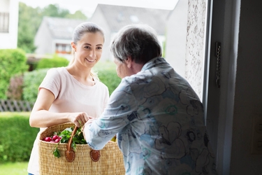 eine junge Frau reicht einer Seniorin einen Korb voller Lebensmittel an der Haustür