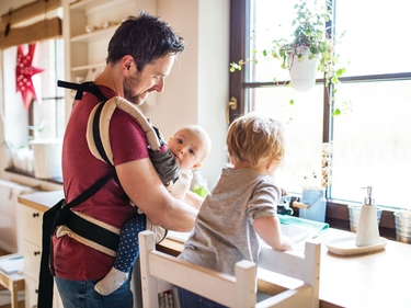 Vater mit einem Baby in einer Tragetasche und einem Kleinkind auf einem Stuhl