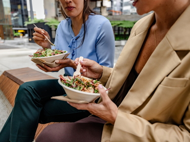 Zwei Personen sitzen draußen und essen ihr Mittagessen aus Schüsseln.