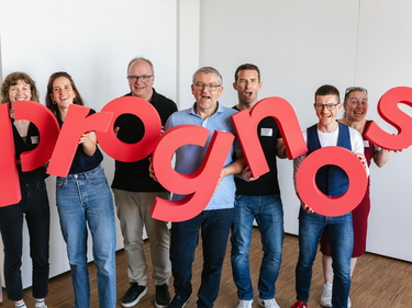 Prognos Mitarbeitende halten rote Buchstaben mit dem neuen Prognos Logo in der Hand