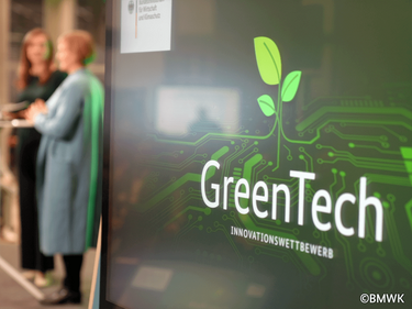 Im Vordergrund das GreenTech Logo aus einer grünen Wand, dahinter zwei Personen, die sich unterhalten