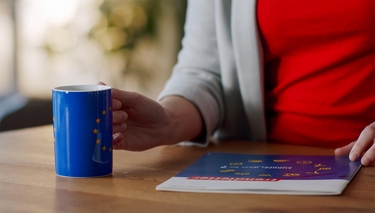 Eine Frau umfasst eine Tasse, auf der die Europaflagge zu sehen ist und schlägt ein Magazin auf