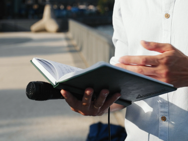 Person im weißen Hemd hält aufgeschlagenes Buch mit Mikrofon, draußen im Sonnenlicht.