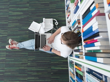 Eine Person sitzt auf dem Boden einer Bibliothek und arbeitet am Laptop