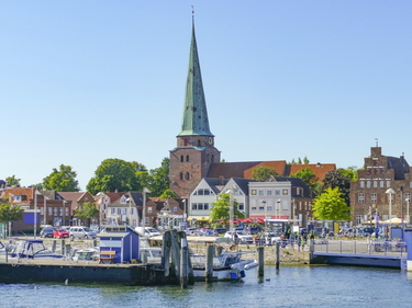 Hafen von Travemünde mit Booten, bunten Häusern und hoher Kirchturmspitze bei Sonnenschein.