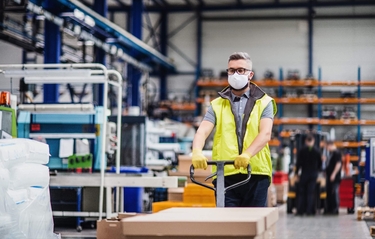 ein junger Mann in Warnweste schiebt Ware auf einem Wagenheber durch eine Produktionshalle