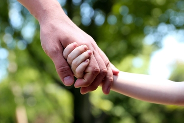 Vater hält die Hand eines kleinen Kindes, im Hintergrund Bäume und Sonnenschein