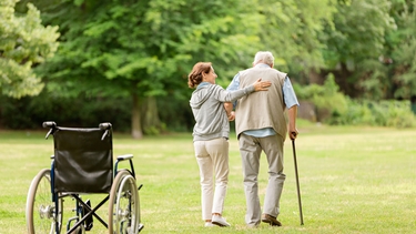 Älterer Mann mit Gehstock läuft mit Pflegekraft auf der Wiese, am Rand steht ein Rollstuhl