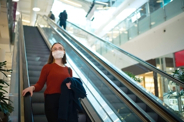 Eine junge Frau mit Mund-Nasen-Schutz steht auf einer Rolltreppe