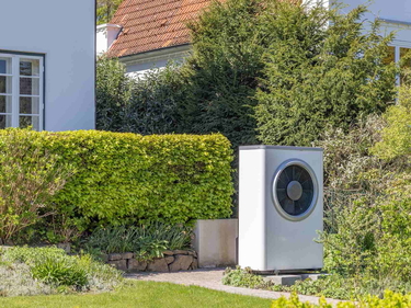 Außeneinheit einer Wärmepumpe im Garten neben Hecke und Hauswand