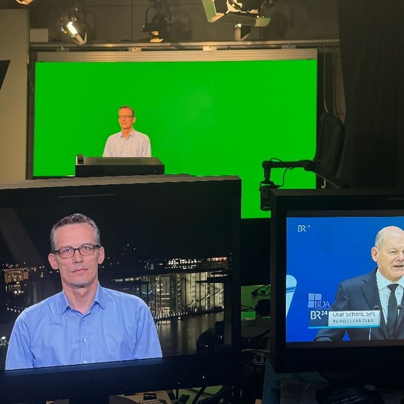 Zwei Aufnahmebildschirme einer TV-Aufzeichnung im Vordergrund, im Hintergrund Sitze Herr Böhmer vor einem Green Screen