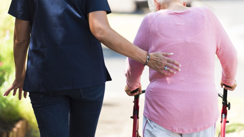 Seniorin geht an einem Rollator, Pflegefachkraft stützt sie 