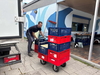 Mann schiebt roten Stapel Kisten auf Rollwagen vor Werkstatt, Transporter daneben