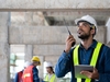 Eine Person auf einer Baustelle mit Warnweste und Helm spricht in ein Walkie-Talkie
