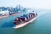 großes Frachtschiff mit Containern beladen fährt an einem Hafen und Hochhäusern dahinter vorbei