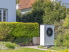 Außeneinheit einer Wärmepumpe im Garten neben Hecke und Hauswand
