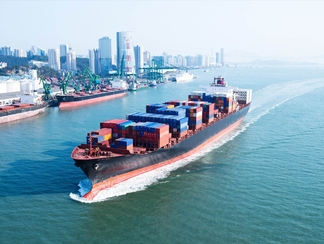 großes Frachtschiff mit Containern beladen fährt an einem Hafen und Hochhäusern dahinter vorbei